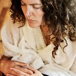 A person with curly hair, wearing a white robe, sits calmly with closed eyes and folded hands.