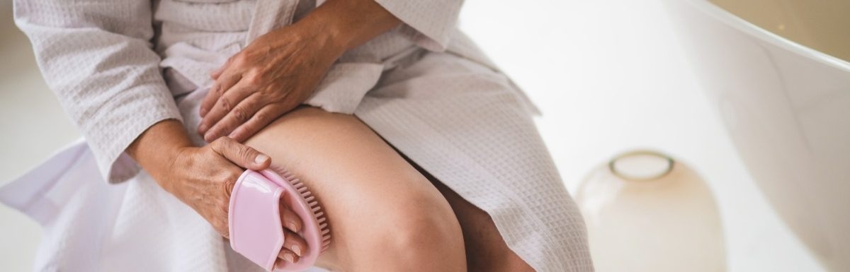 Person in a bathrobe practicing drybrushing on their leg while sitting near a bathtub.