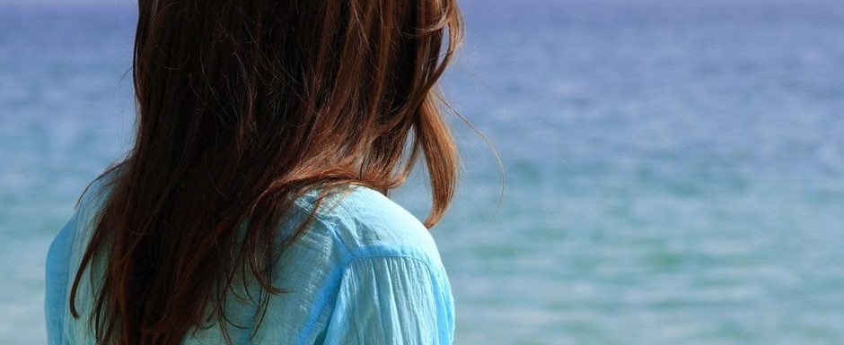 A woman wearing a blue shirt looking at the ocean, reflecting on past experiences.