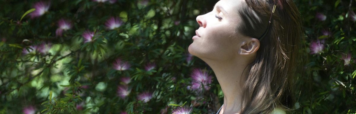 A woman with sunglasses on her head stands peacefully, eyes closed, facing the sun. Embraced by green foliage and pink flowers, she takes a moment to savor the benefits of nasal breathing in nature's embrace.