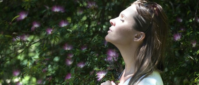 A woman with sunglasses on her head stands peacefully, eyes closed, facing the sun. Embraced by green foliage and pink flowers, she takes a moment to savor the benefits of nasal breathing in nature's embrace.
