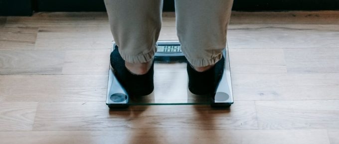 A person stands on a digital glass scale, potentially influenced by hormone leptin, wearing black socks and beige pants on a wooden floor.