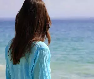 A woman wearing a blue shirt looking at the ocean, reflecting on past experiences.