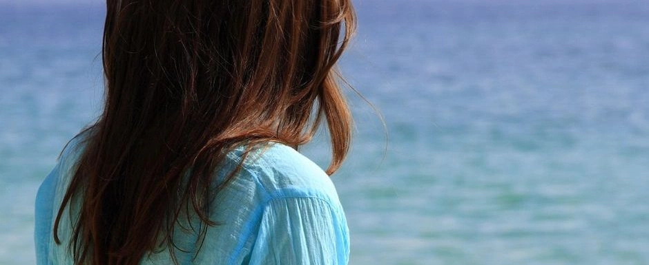 A woman wearing a blue shirt looking at the ocean, reflecting on past experiences.