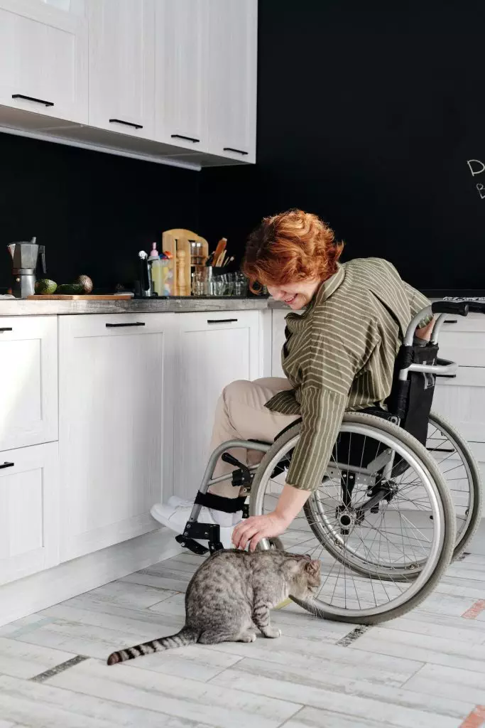 A woman in a wheelchair finds comfort in petting a cat while practicing holistic health tips in her kitchen.