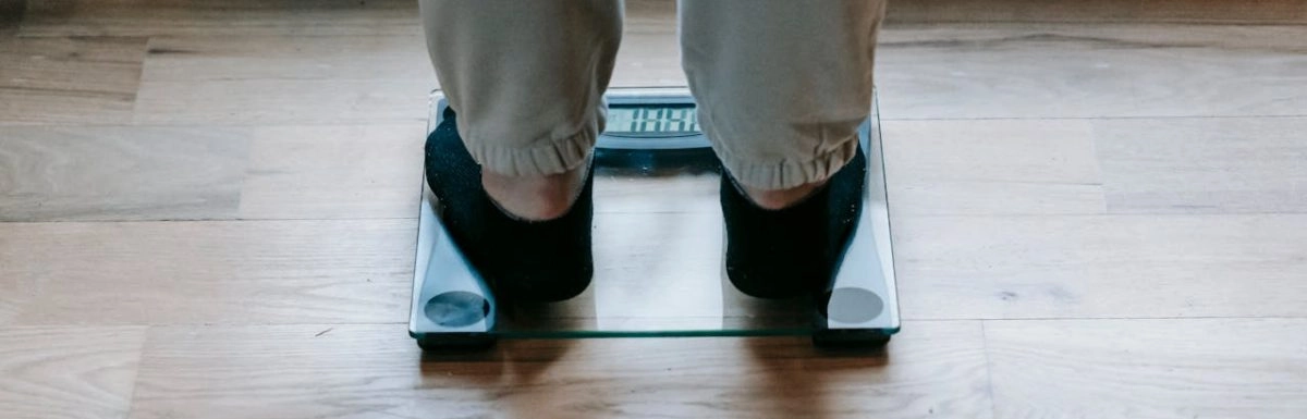A person stands on a digital glass scale, potentially influenced by hormone leptin, wearing black socks and beige pants on a wooden floor.
