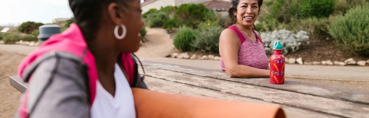 Two women are sitting on a picnic table outdoors, one holding a yoga mat and the other a water bottle. Engaged in deep conversation beneath the gentle sway of trees, they find balance and grounding akin to the Muladhara Root chakra with a building and greenery in the background.