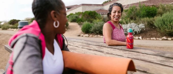 Two women are sitting on a picnic table outdoors, one holding a yoga mat and the other a water bottle. Engaged in deep conversation beneath the gentle sway of trees, they find balance and grounding akin to the Muladhara Root chakra with a building and greenery in the background.