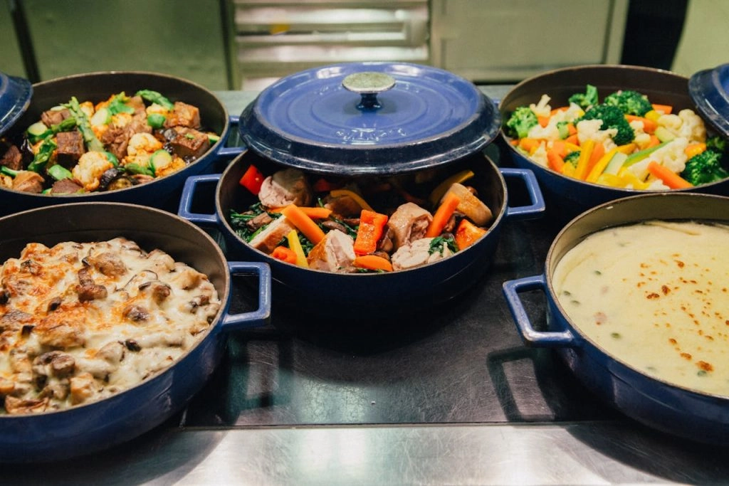 Five blue pots filled with various dishes, including meat, vegetables, and a creamy sauce, displayed on a countertop.