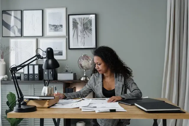 A stressed out woman sits at a desk reviewing papers, surrounded by office supplies, with framed art on the wall.