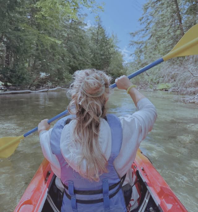A woman in a kayak, moving with the flow of the river, surrounded by lush green trees under a vibrant blue sky.