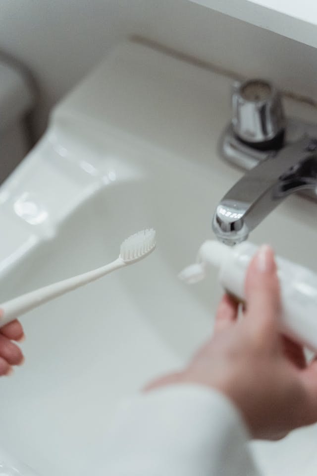 A person applying fluoride toothpaste onto a toothbrush over a white sink with a chrome faucet.